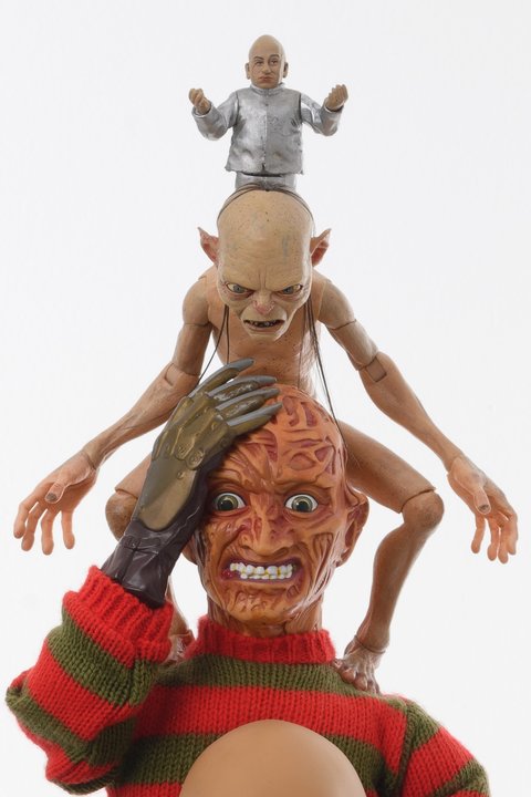Close-up image of a sculpture from the exhibition featuring a Freddy Krueger character figurine with a red and brown striped shirt, and bloody raw muscle face. It is holding a brown gloved hand up to its forehead. A Gollum character figurine is seated atop the Freddy Krueger figurine. The figurine looks down onto the figure below with its naked body and arms stretched out to the side. Atop Gollum, sits a Dr. Evil character figurine complete with a bald head, silver top and pants. The arms are outstretched as it sits atop the other two figurines. 