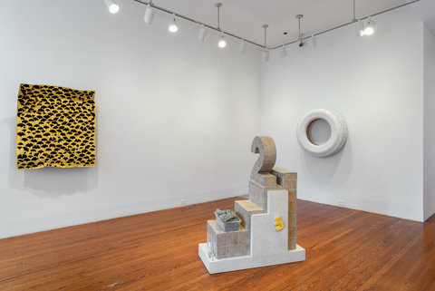 Installation view of the exhibition featuring three artworks. On the left hand side there is a yellow rectangle mounted on the wall made of cardboard. It is covered in stamps of black ginko leaves. On the ground in the middle is a tile statue of the number 2. Behind the statue, mounted on the wall on the right is a white tire made of fiberglass with very light orange ginko leaves stamped on to it.  