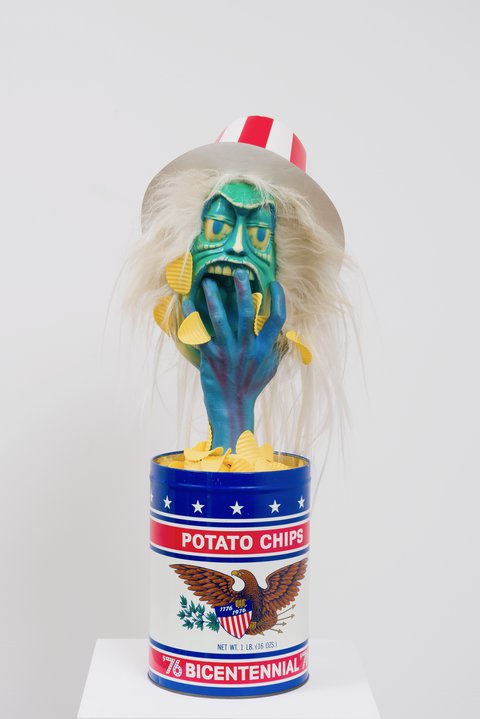 Close-up image of a sculpture of a potato chip tin with the words "POTATO CHIPS, 76 BICENTENNIAL' written in typeface across the top and bottom. In between sits an eagle holding an emblem of red and white stripes and a branch. Atop the potato chip tin, which is open, with fake potato chips, emerges a blue hand holding that is stuffing potato chips into the mouth of a green, zombie-like Uncle Sam figurine. The figure wears a red and white striped hat and has strands of white and grey hair flying out from under its hat. The figure has droopy scary eyes and teal skin. 