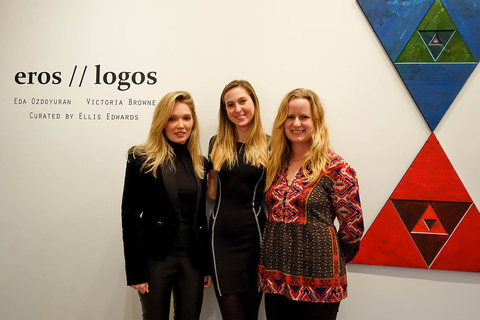 View of three visitors in the gallery in front of a vinyl wall text that states, "EROS // LOGOS, VICTORIA BROWNE AND EDE OZDOYURAN, CURATED BY ELLIS EDWARDS." To their right, two triangles sit on top of one another, point to point. The top triangle, is blue and the bottom is red. 