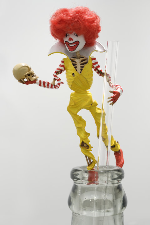Close-up image of a skeleton figure standing on the mouth of a clear, glass bottle. It is holding onto the straw that emerges from the bottle. The figure is a skeleton dressed up as a Ronald McDonal character figurine. With red shoes, a yellow jumpsuit, white and red striped long sleeve shirt, and a big white color. The figure holds a small skull in its right hand. The figure has a white painted face, with a red nose, and big red painted clown lips. The figure has large red fluffy hair. 