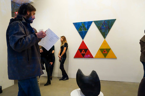 Installation view with visitors frequenting the exhibition. A visitor stands in the foreground next to a pedestal with a black, abstract sculpture sitting atop it. In the background, four triangles are organized together in rows of two by two, the top triangles points touch the bottom triangles points and tehy are colored blue, red, and yellow. 