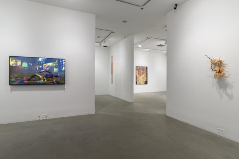 Gallery installation view featuring two walls leading into two other gallery rooms. On the left wall, a mounted TV with a video still featuring bright colors and lights with dancing animated, computer animated images. On the right is a tannish wood sculpture featuring gourds and wood that spikes out around the center.