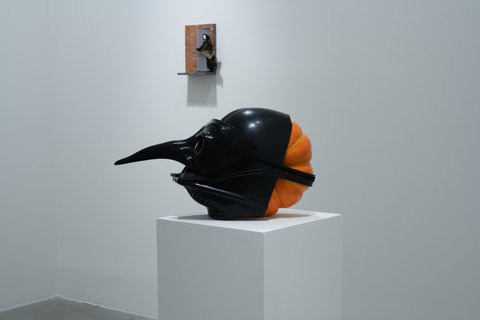 Installation view of the exhibition featuring a orange pumpkin with a black mask on it. The black mask has a long pointed nose, like a crow. The sculpture sits on a white pedestal. In the background of the image, there is a artwork on the wall featuring a a skeletal figure dressed in a black cloak, mid-run as it pushes open a grey door against a brick wall. The figure holds a small piece of art in a gold frame.  