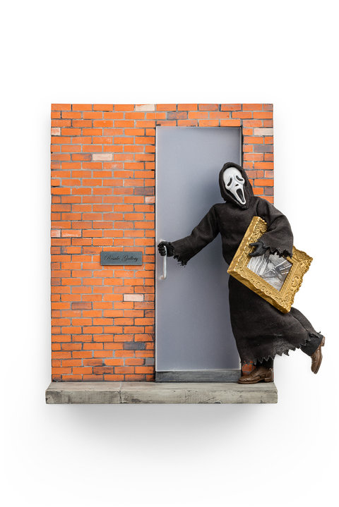 Close-up image of a sculpture in the exhibition, hung on a wall, featuring the grim reaper, a skeletal figure dressed in a black cloak, mid-run as it pushes open a grey door against a brick wall. The figure holds a small piece of art in a gold frame. 