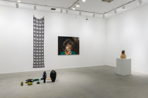 Gallery installation view featuring a wire mesh rectangle hanging from the ceiling. Below it sits five separate ceramic pots in order from smallest to largest. Next to the hanging piece, on the back wall in the background is a figural painting of a figure with an Afro hairstyle. The figure wears a green shirt, although only the figures neck and head and a small portion of their shoulders are included in the image. On the right is a pedestal with what looks like a piece of blown glass. It is neutral in color, like a beige or grey. 