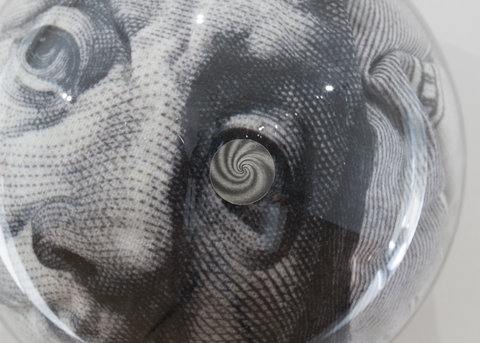 Up-close image of a sculpture in the installation. There is a glass orb that magnifies an image of a president from a paper piece of money. Only the man's left eye, right eye, and nose are shown on left facing profile. The man's right eye is replaced with a spiral design. 