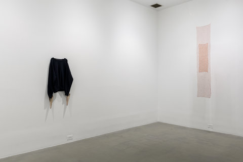 Gallery installation view of two walls. On the left wall, there is a black sweatshirt hung up at the corner shoulders. On the right wall, hangs from the ceiling, a copper knit-mesh sculpture. It is suspended about two to three feet off the ground and  hangs down from the ceiling at about 70 inches or six feet tall.