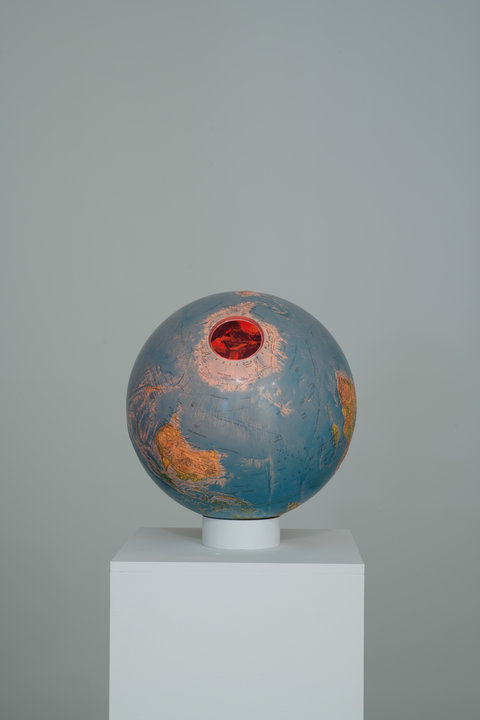 Image of a sculpture in the exhibition of an orange and grey-ish blue globe. The globe sits on a pedestal. There is a circular hole on the top of the globe. 