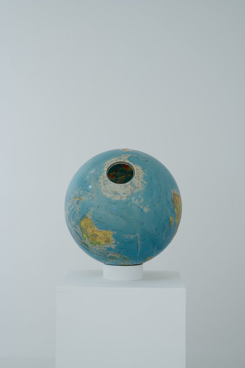 Image of a sculpture in the exhibition of a green and blue globe. The globe sits on a pedestal at an angle. There is a circular hole on top of the globe. 