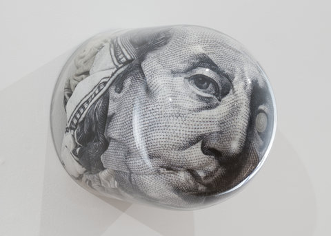 Image of sculpture made from a glass orb that magnifies a close up picture of an old man, most likely a president on a piece of paper money. It is in black and white and only a part of the face is magnified. 