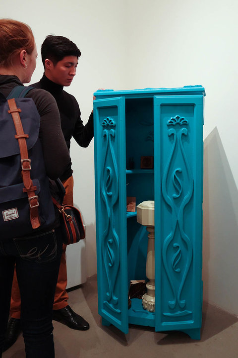 Installation image from the exhibition featuring a blue cabinet. Two figures, visitors of the exhibition, stand near the cabinet and peer into it. The cabinet sits on the floor of the gallery and is ajar. 