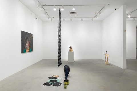 Installation view of the exhibition featuring four works. The image shows three walls and the center of the room in which there is a hanging sculptural piece. On the left wall, there is a painting of a woman with an brown afro and green background. The image is up-close and only the figures head is shown. On the back wall, on a pedestal sits a abstract glass sculpture. On the right side there is a small tan wooden table with a diet coke can sitting atop it. In the center of the room, there is a hanging metal piece, from side view. It is black and hangs down above various ceramic pieces that are seated on the floor. 