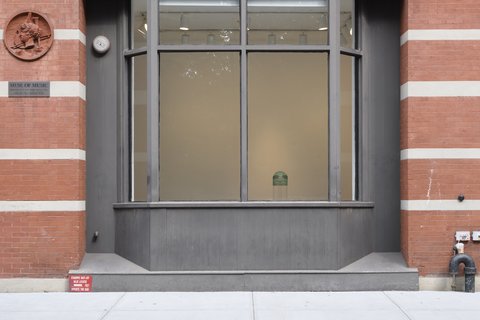 Outside view of the gallery space. There is one large window with two panels. From the outside, it appears to be only white walls. On either side of the large window pane, are red bricks of the building evenly and intermittently striped with white brick. 