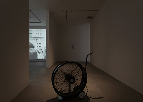A black fan on wheels with a handle in a dark corner of the gallery. Shot from behind, a room with a projection on the wall can be seen in the distance.