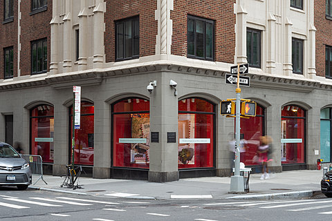 The corner of a building with six windows seen from street-level. People walk in front of ground floor windows filled with various things against red backgrounds.