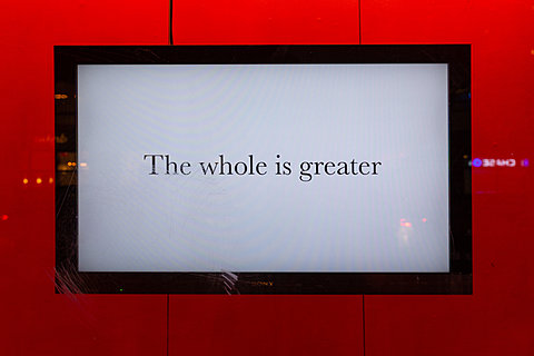 A television monitor displaying the text "The whole is greater" hangs against a red background.