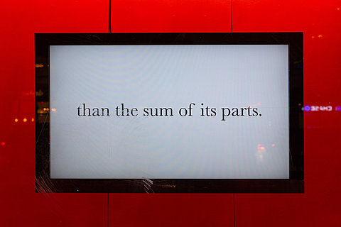 A television monitor displaying the text "than the sum of its parts." hangs against a red background.
