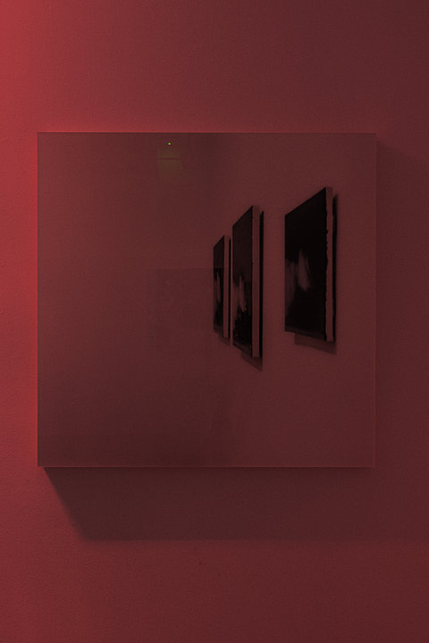 In a room filled with red light, there is a print on the wall showing the image of two artworks hanging on wall, taken from an oblique angle.